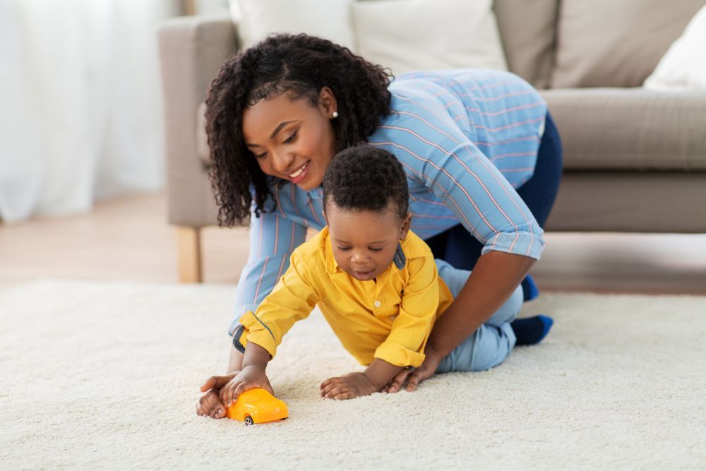 Woman kneeling with child playing with a toy car on a carpet.