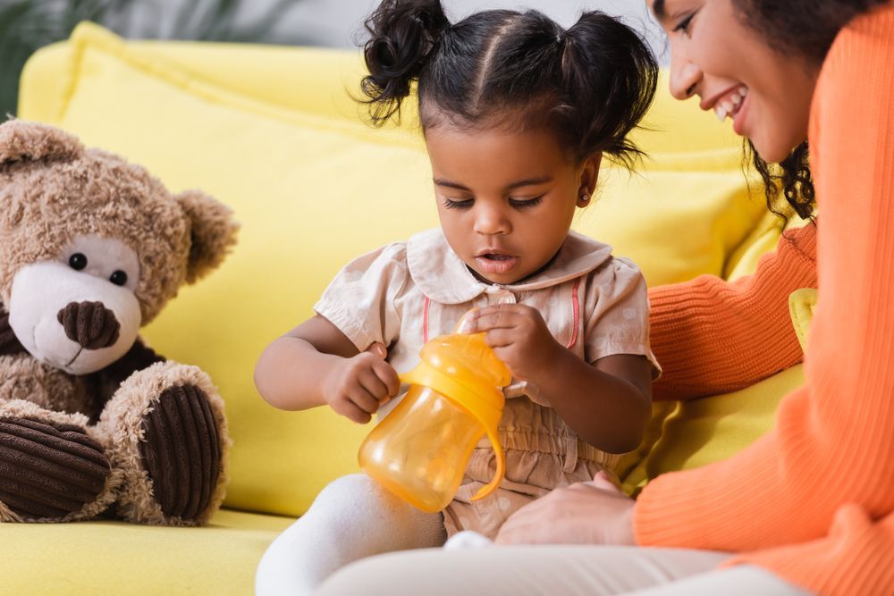 Child seated with bottle, alongside adult and teddy bear on a yellow couch.