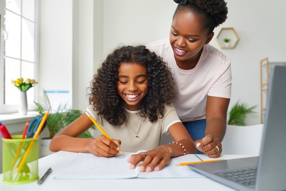Woman helping a child with homework at a desk with a laptop and pen. Both smiling.