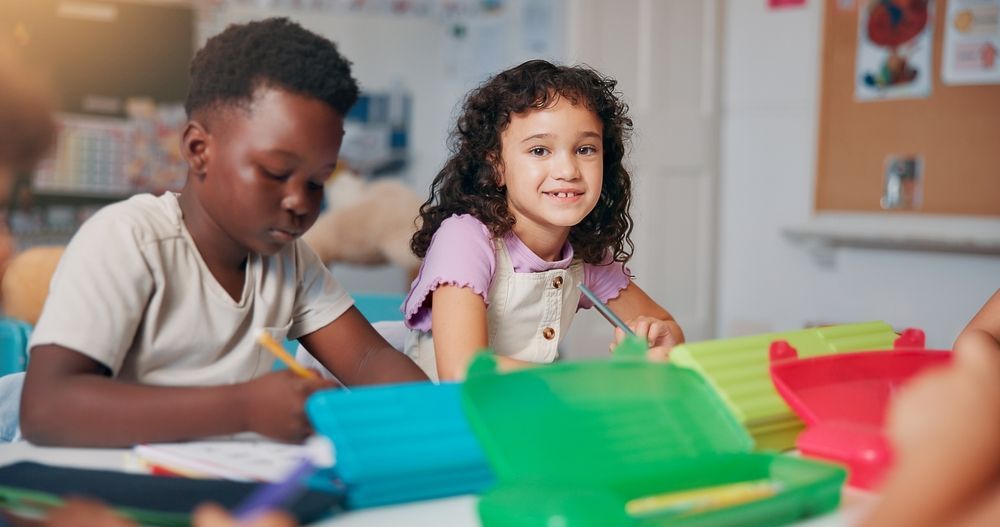 Two children at a desk in a classroom; girl smiles at camera, boy writes. Green, red pencil cases.
