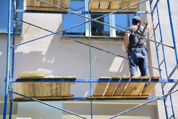 Man painting exterior wall, standing on scaffolding next to a window; blue sky.