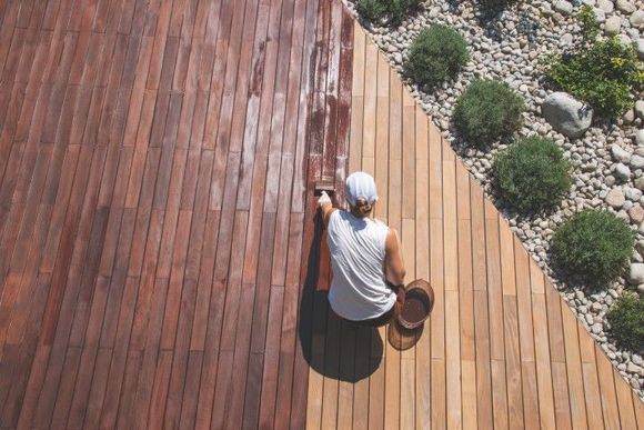 Person staining wooden deck; half stained dark brown, other half unfinished.