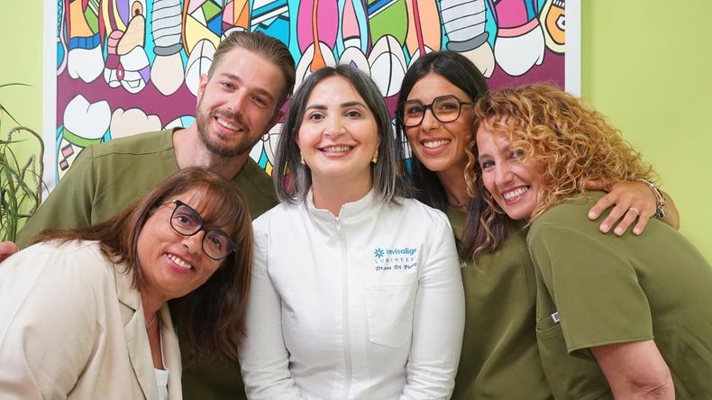 Cinque persone sorridenti posano insieme in un ambiente interno, indossando magliette identiche verdi e bianche, davanti a un murale colorato.