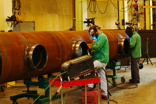 Two men are welding a large pipe in a factory.