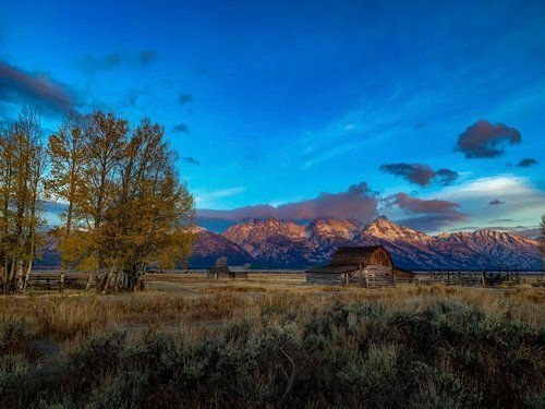 Autumn field with barn and trees under a blue sky, backed by sunlit mountains at sunset