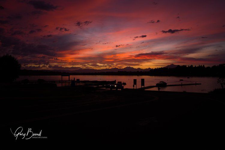 Fiery red-orange sunset over calm waterfront with silhouetted dock and distant mountains
