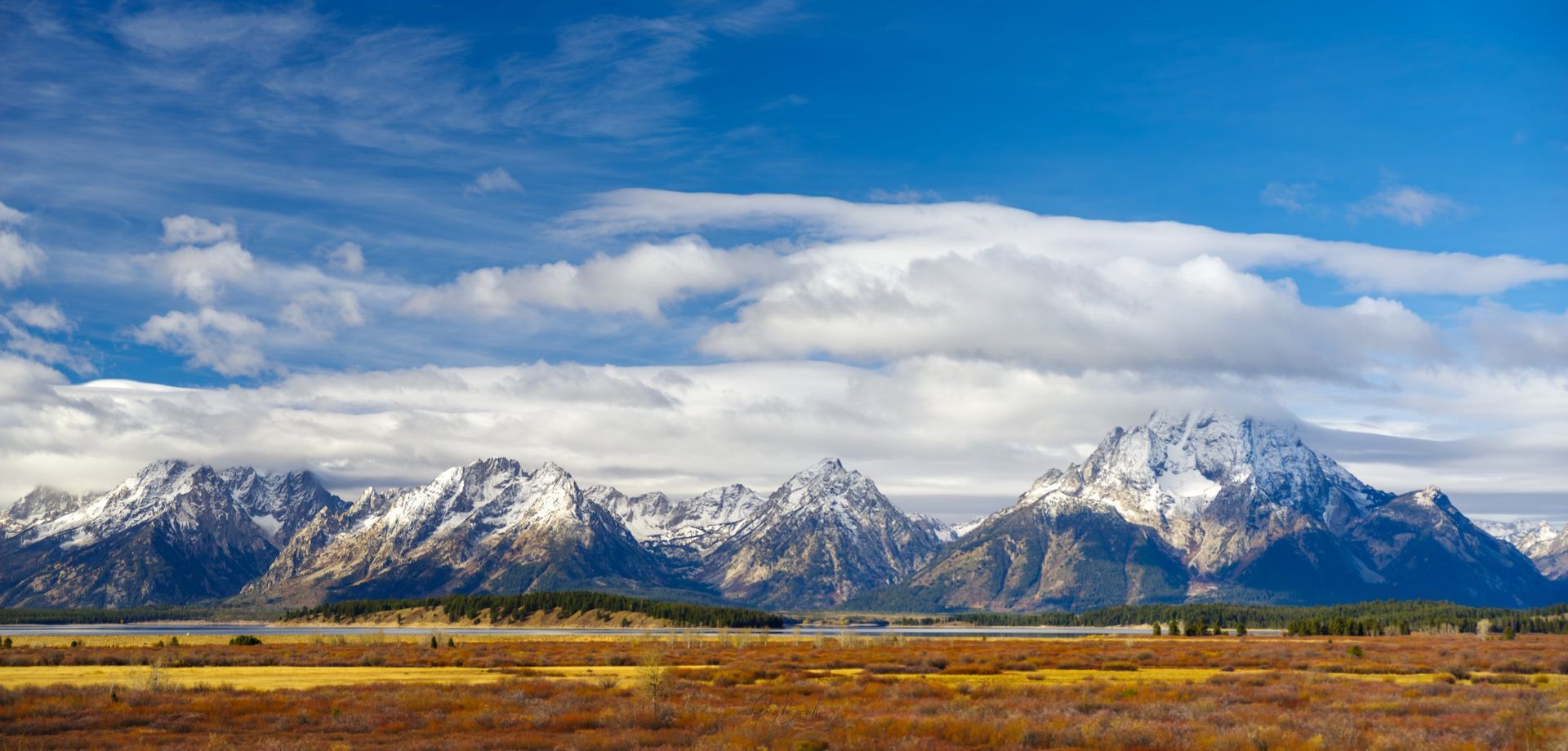Snow-capped mountain range under a cloudy blue sky above a golden field