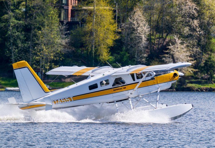 Seaplane skimming across a lake, creating a white spray with forested shoreline in the background