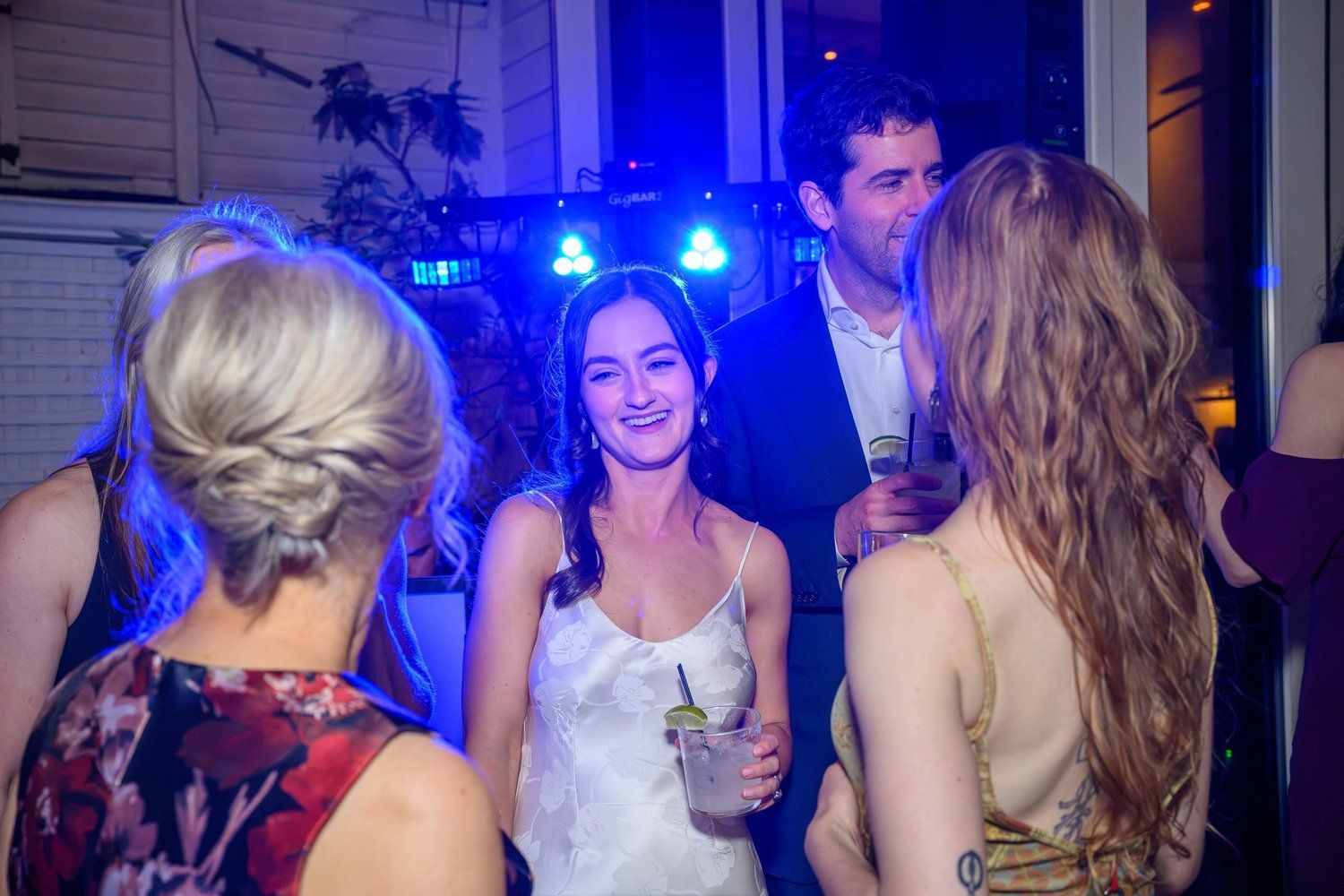 Bride smiling with drink at a lively party, surrounded by guests and blue-purple lights