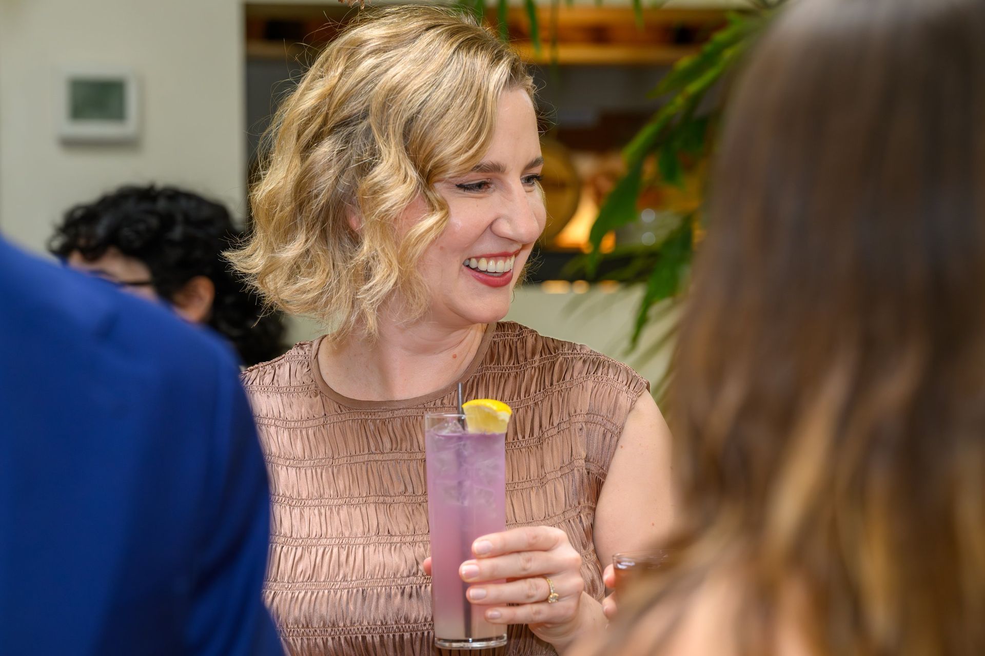 Woman smiling at a social gathering, holding a tall pink drink with a lemon garnish.