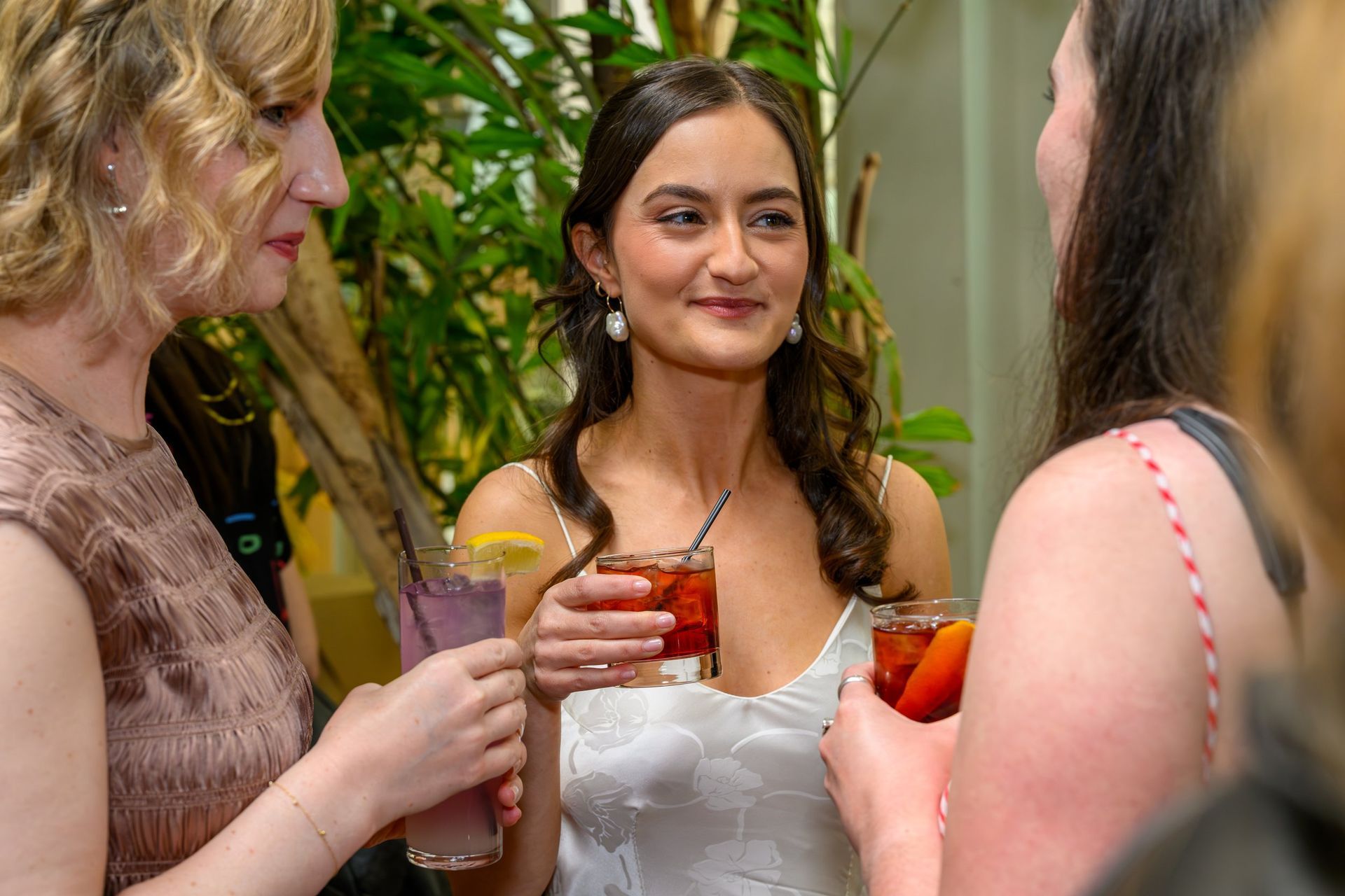 Three people chatting at a party, holding colorful drinks and smiling in a decorated indoor setting
