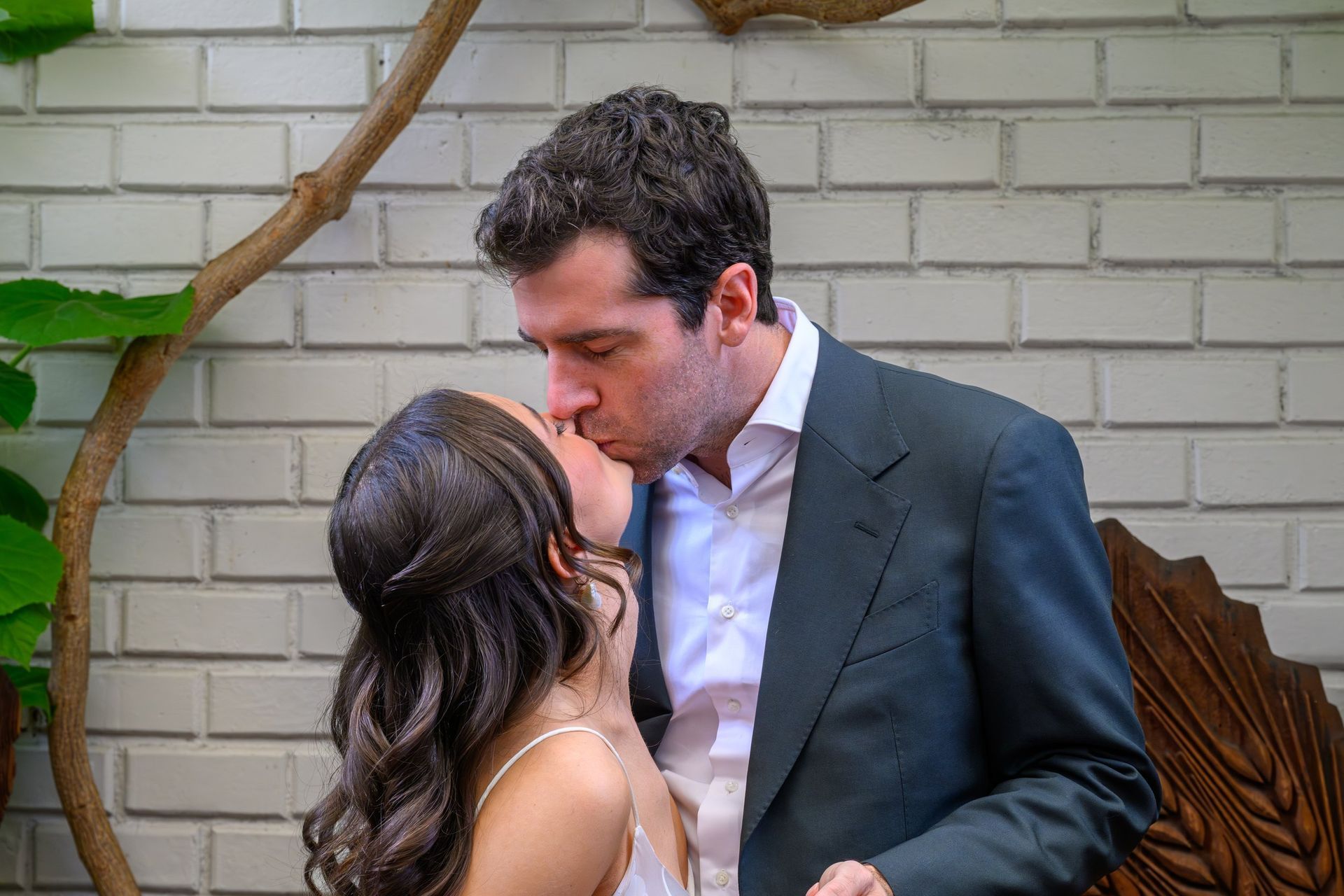 Couple kissing outdoors beside a white brick wall, with greenery and a wooden chair in the background.