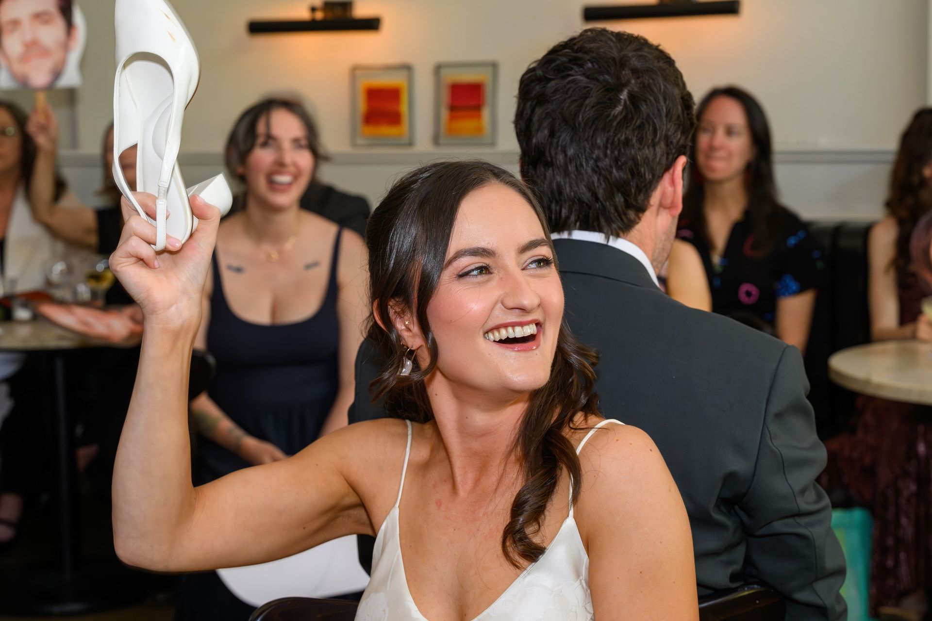 Woman in white dress smiling and holding up a shoe at a lively indoor party