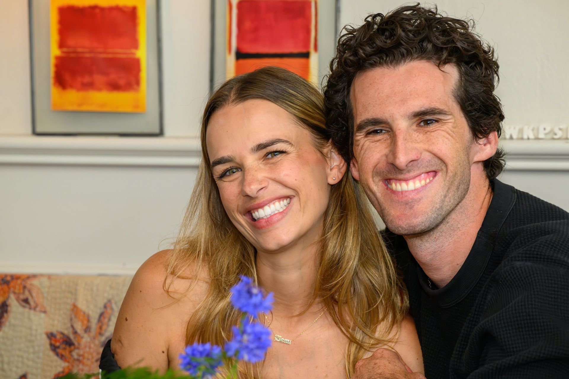 Smiling couple posing indoors on a couch with framed art and purple flowers in the foreground