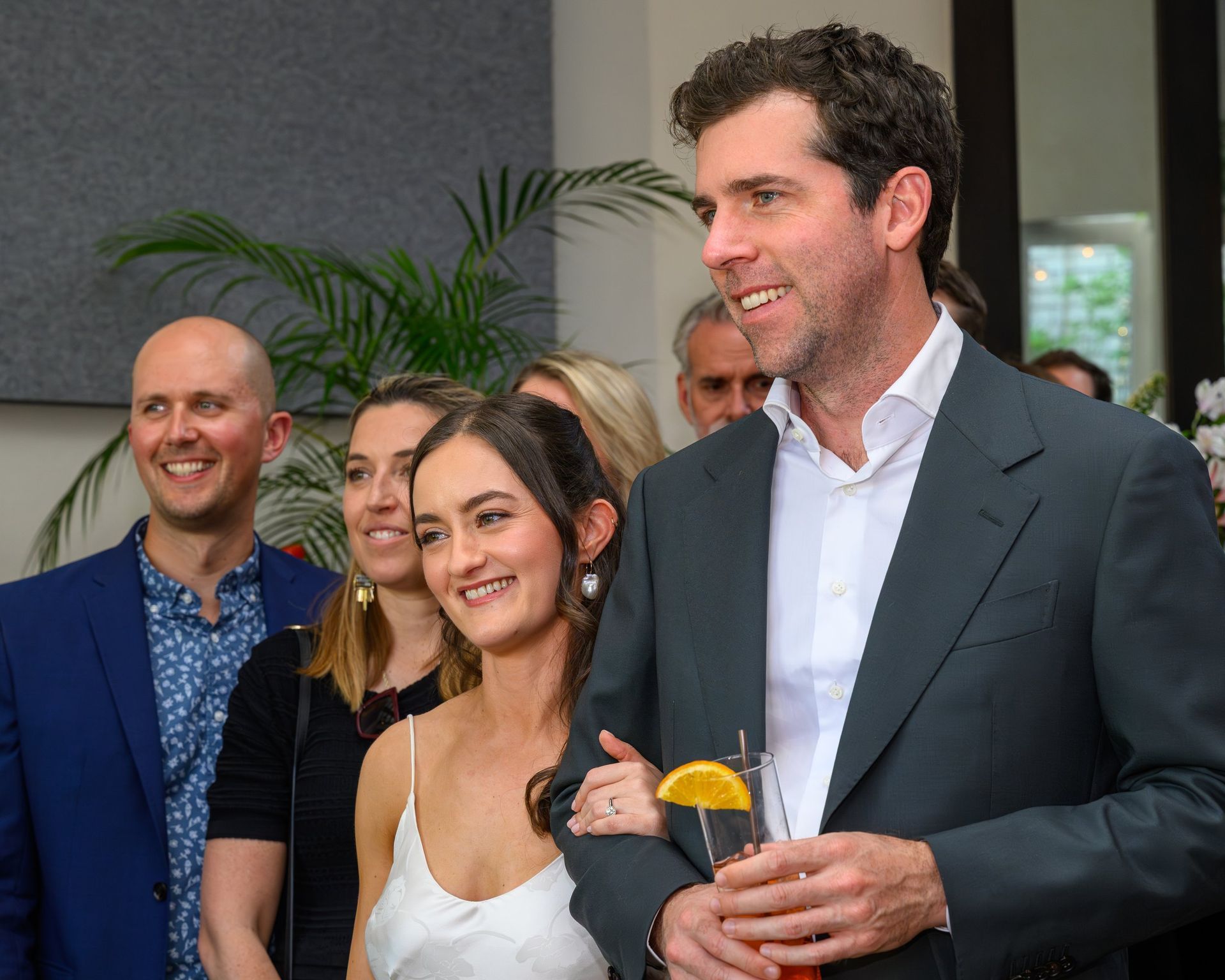 Group of people at an indoor event, smiling and holding drinks, with a tall man in a dark suit in front.