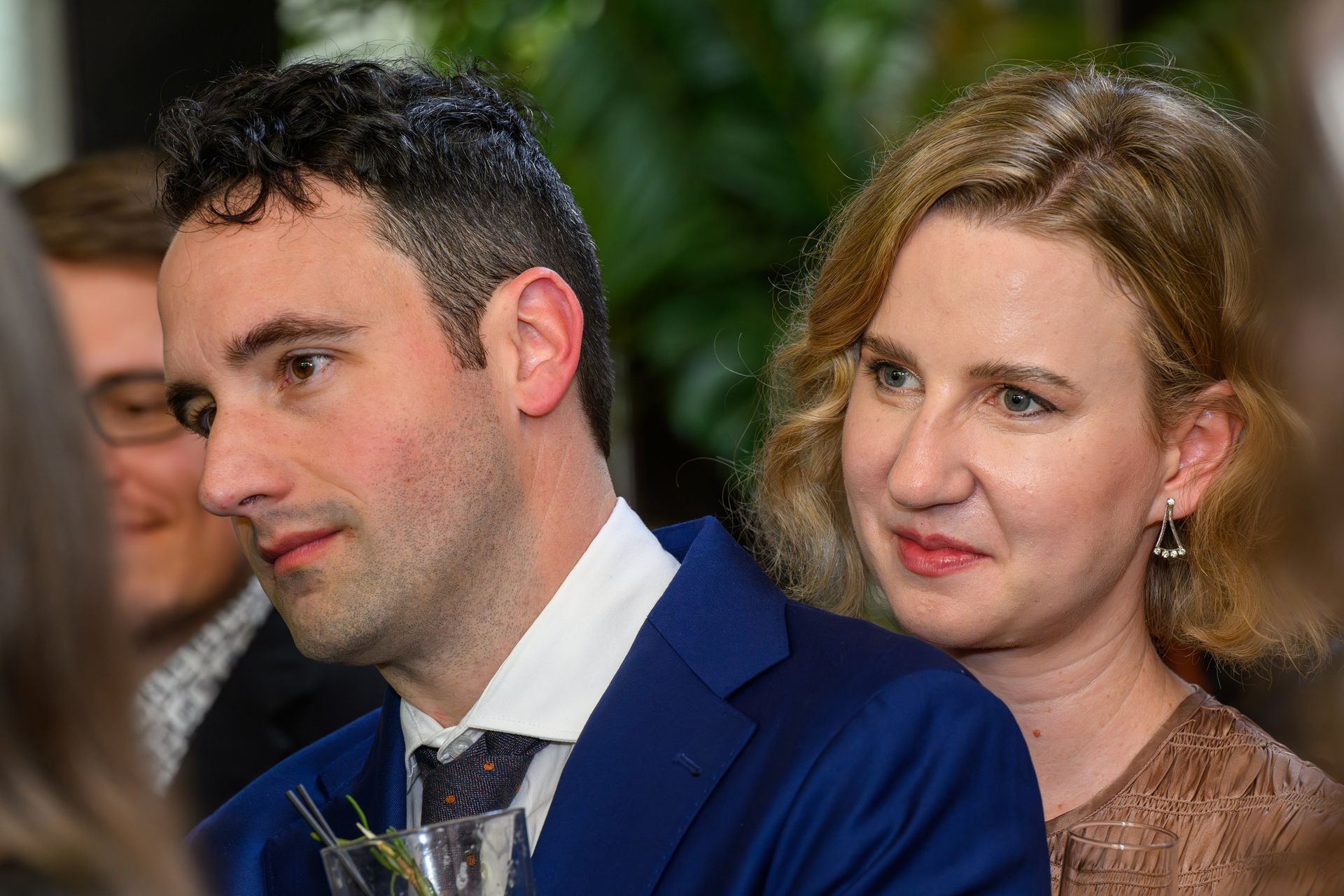 Two people seated close together at an indoor event, one in a blue suit and the other in a beige dress, both looking forward
