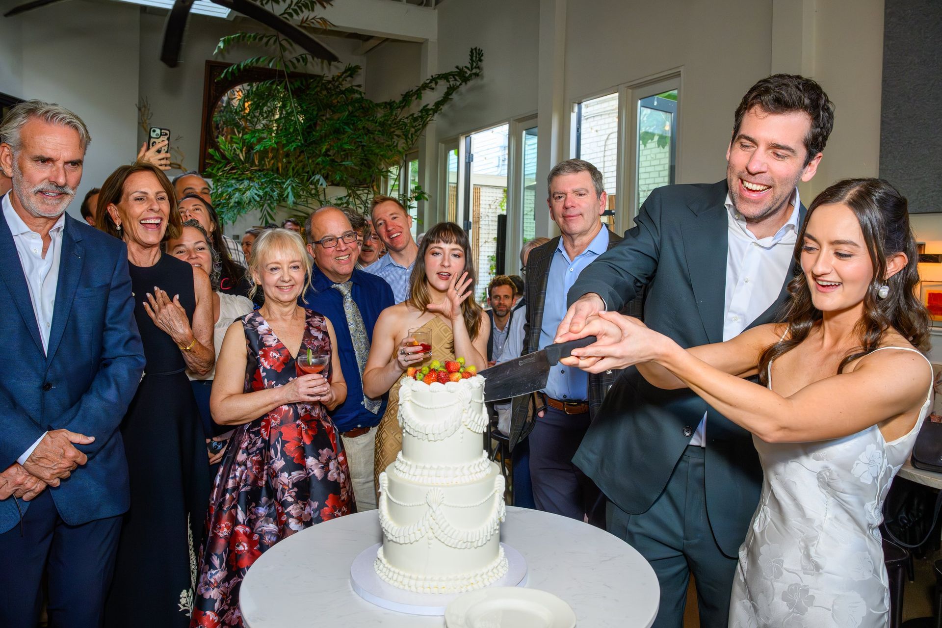 Couple cutting a white wedding cake as guests smile and watch at an indoor reception