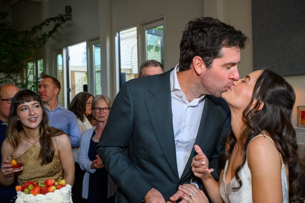A couple kissing at an indoor celebration, with guests watching nearby.