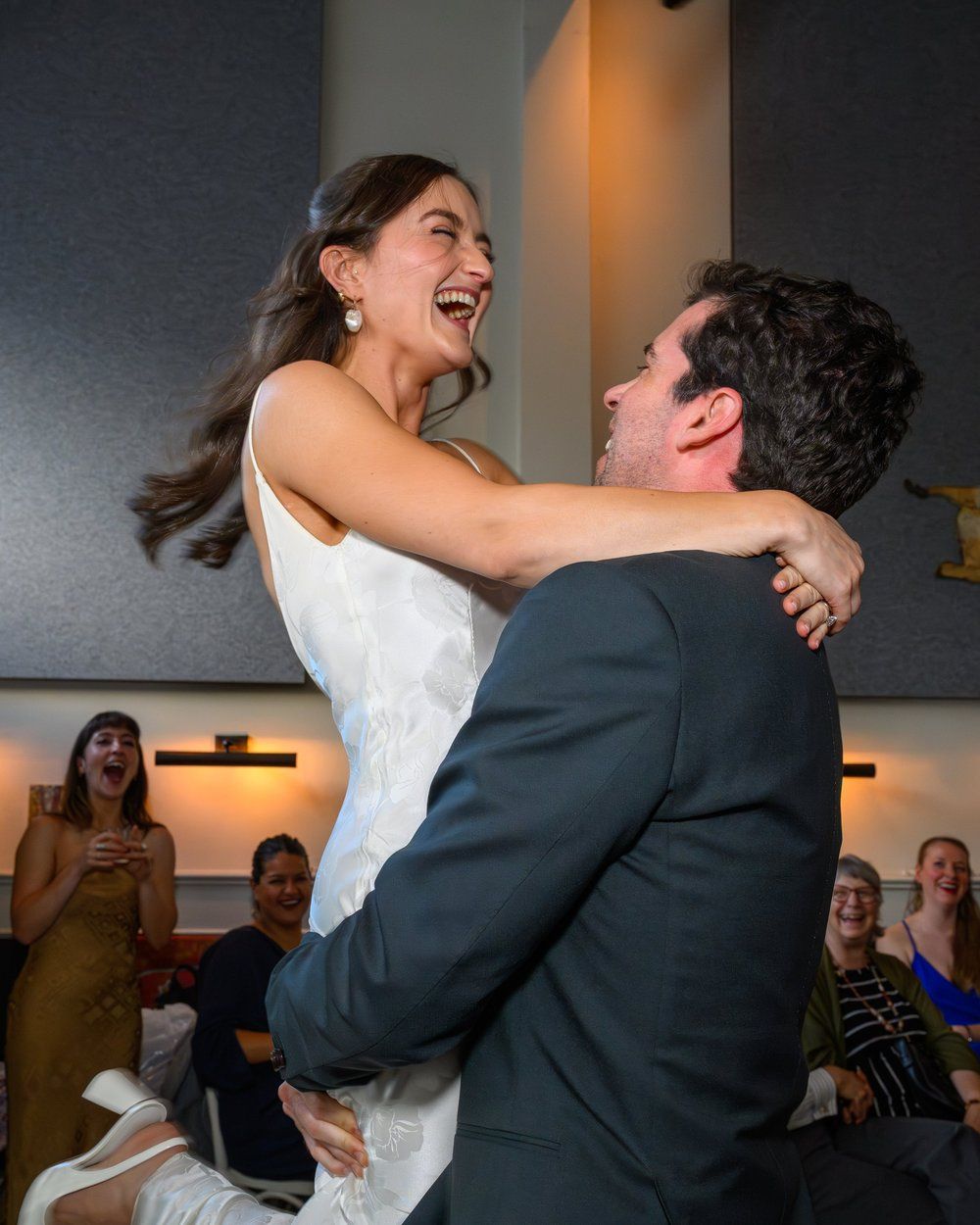 Woman in a white dress and man in a dark suit smiling and embracing at an indoor celebration.