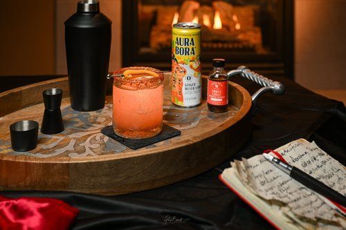 Cocktail setup on a tray with a glass, shaker, bottle, can, jigger, and notebook on a dark table.