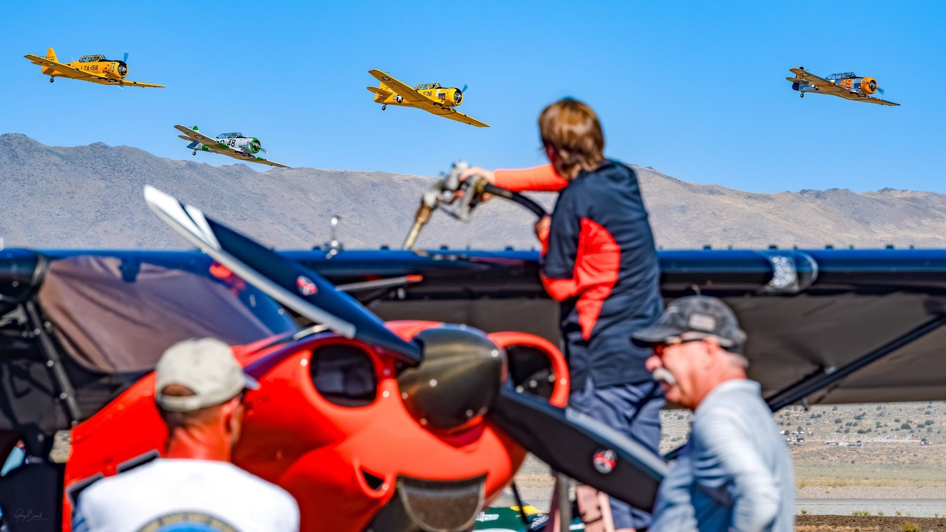 Small aircraft on tarmac with people working nearby as several planes fly overhead against mountains