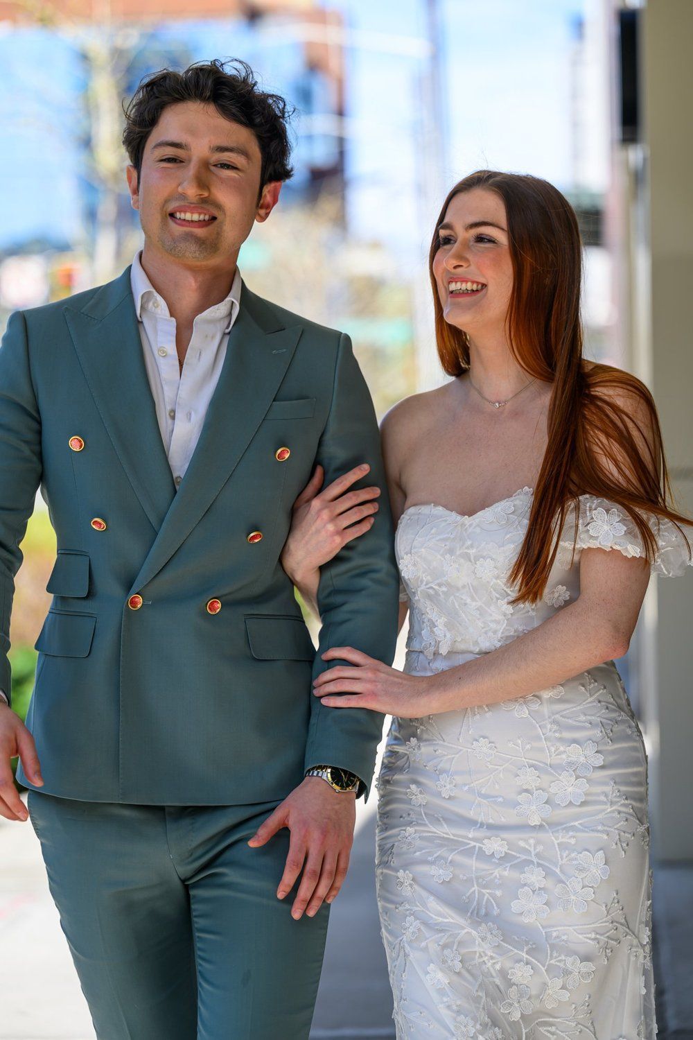 A smiling couple in formal attire walks outdoors, with the woman in a white dress and the man in a green suit.