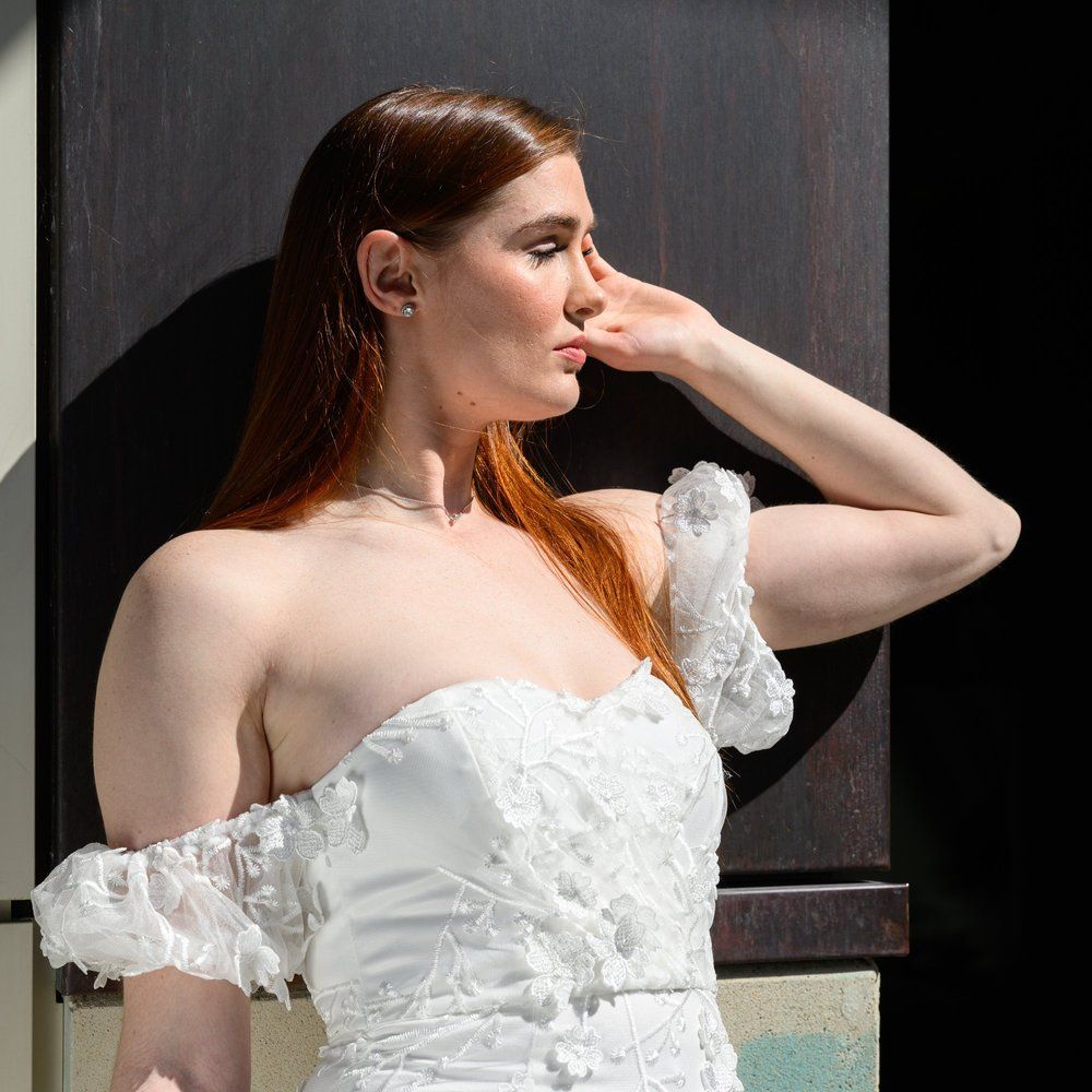 Woman in a white off-shoulder dress posing in profile against a dark wall, hand raised to her face