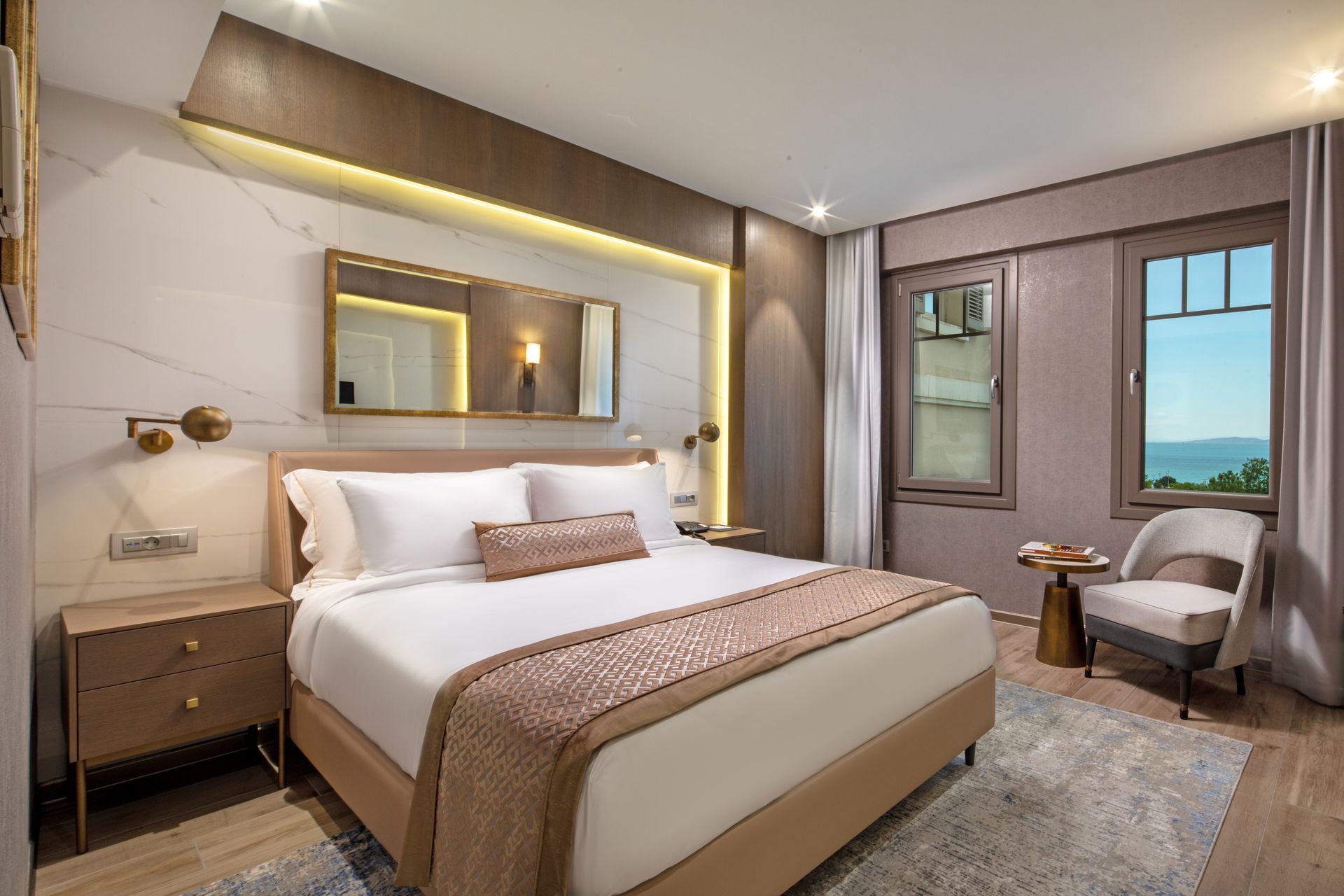 Modern hotel bedroom with a large bed, warm lighting, armchair, and window with a sea view