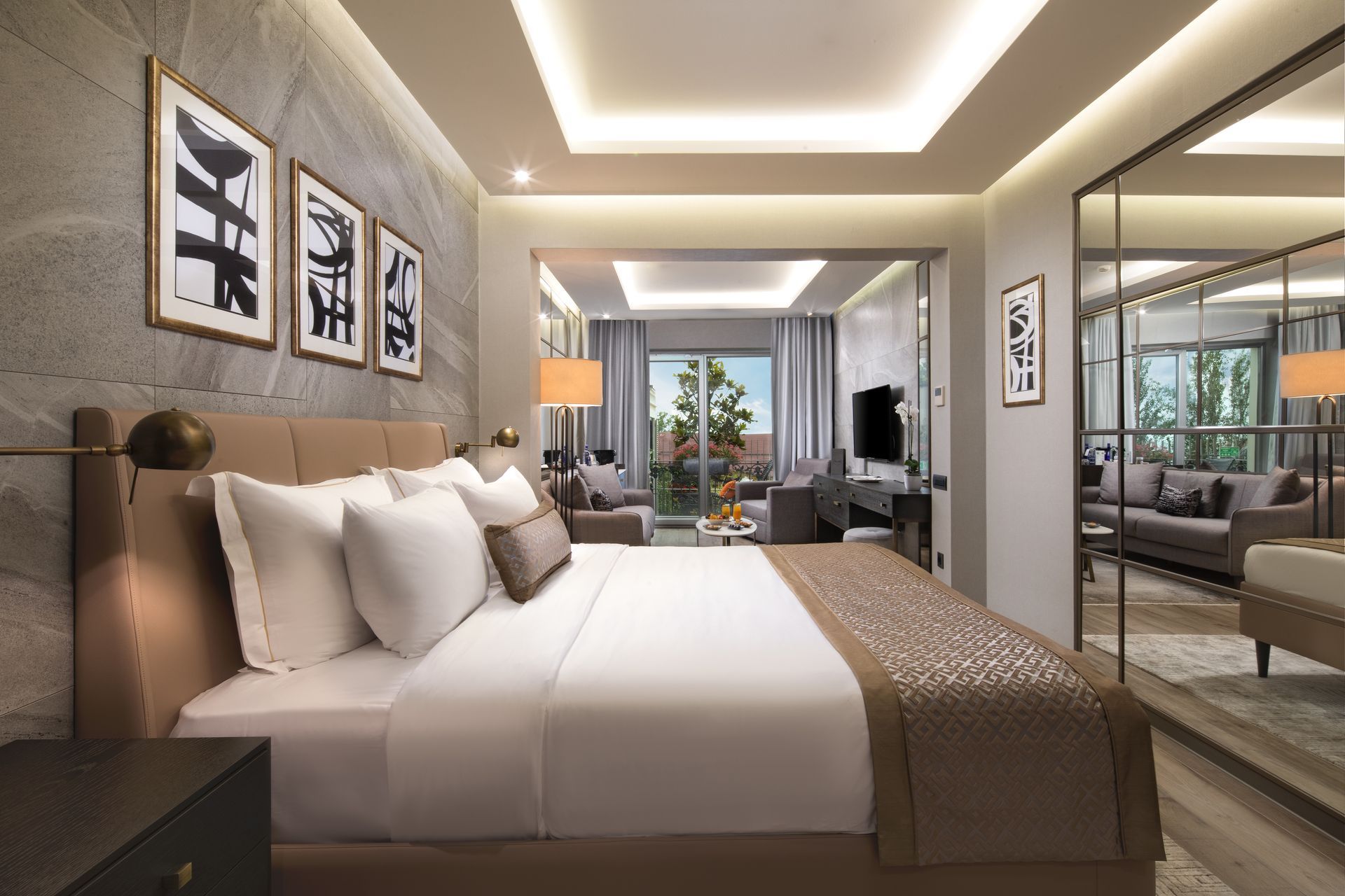 Modern bedroom with a large bed, warm lighting, wall art, and a mirror reflecting a windowed sitting area