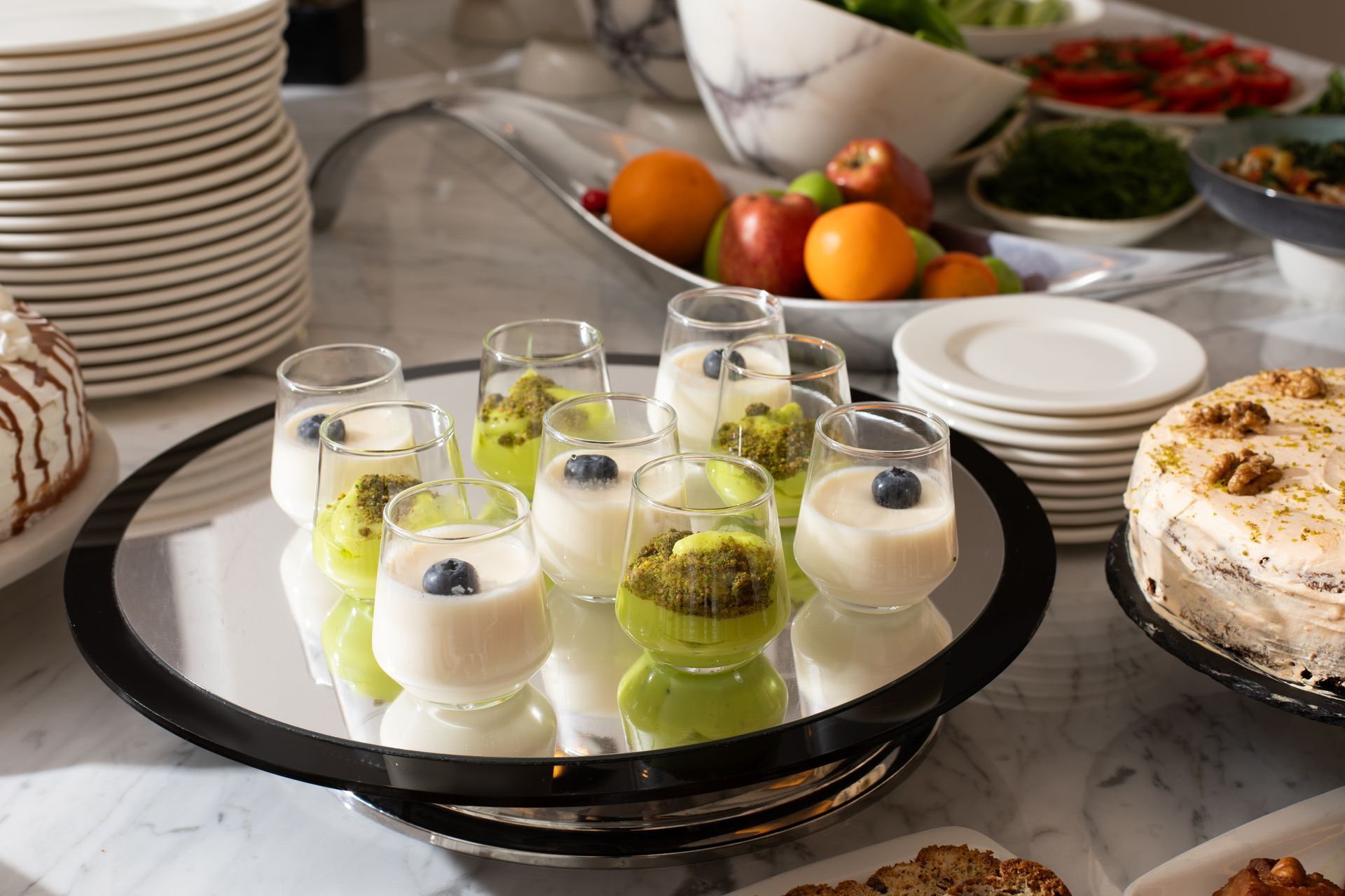 Dessert platter with small white and green mousse cups on a dining table with plates, fruit, and cake