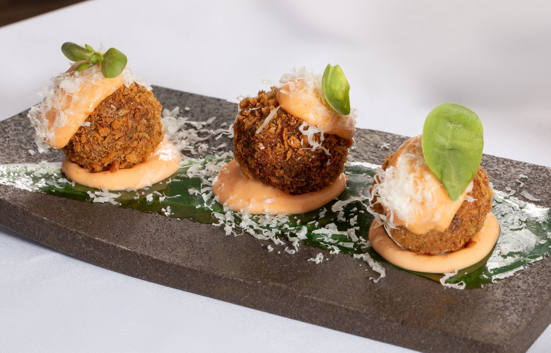 Three plated gourmet appetizers on a slate board with greens and grated cheese, on a white tablecloth