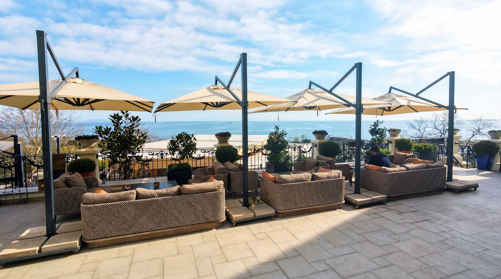 Outdoor seaside café seating with beige umbrellas, stone planters, and ocean view under a blue sky