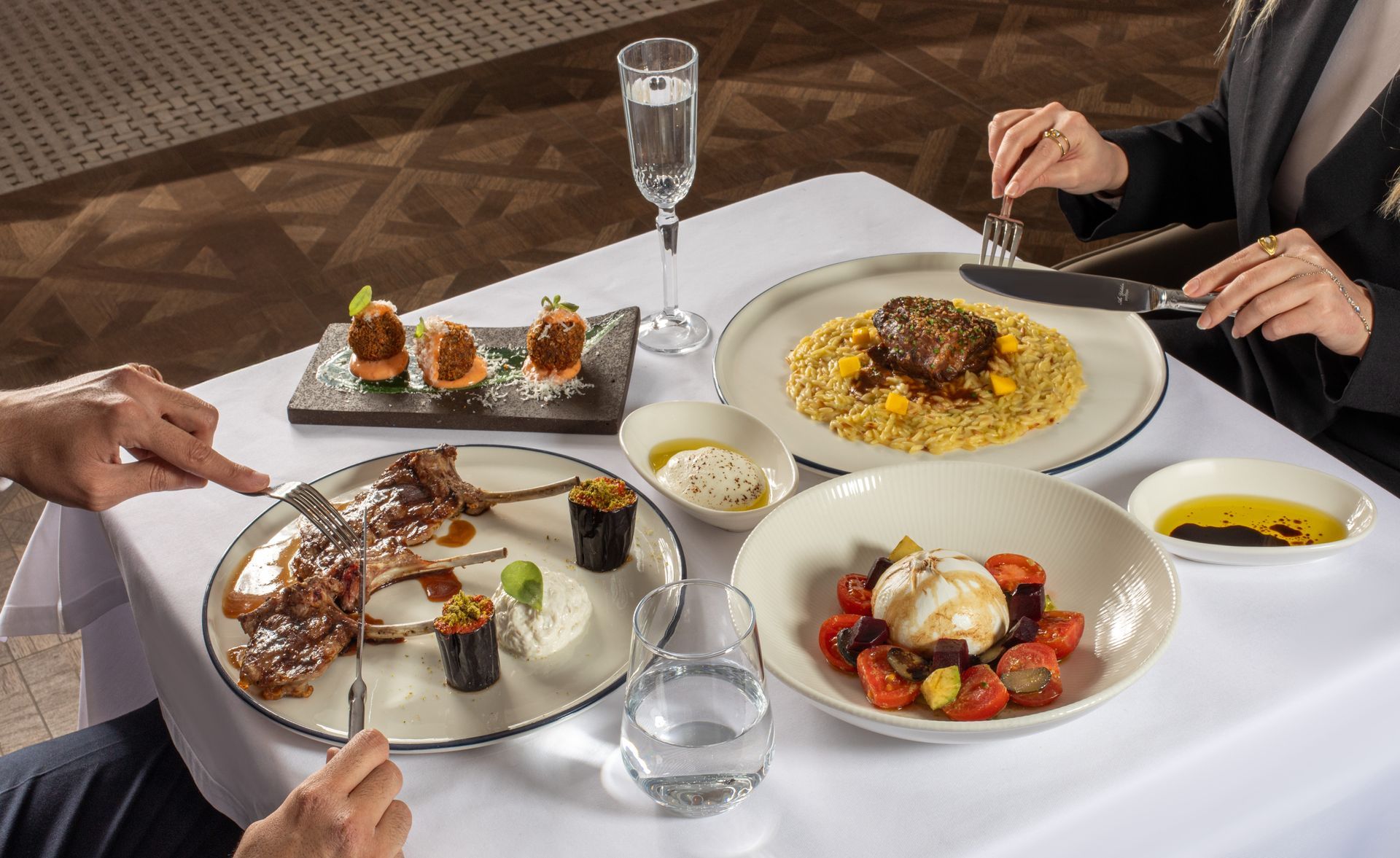Dining table with plated gourmet dishes and drinks, people reaching to eat in a restaurant setting