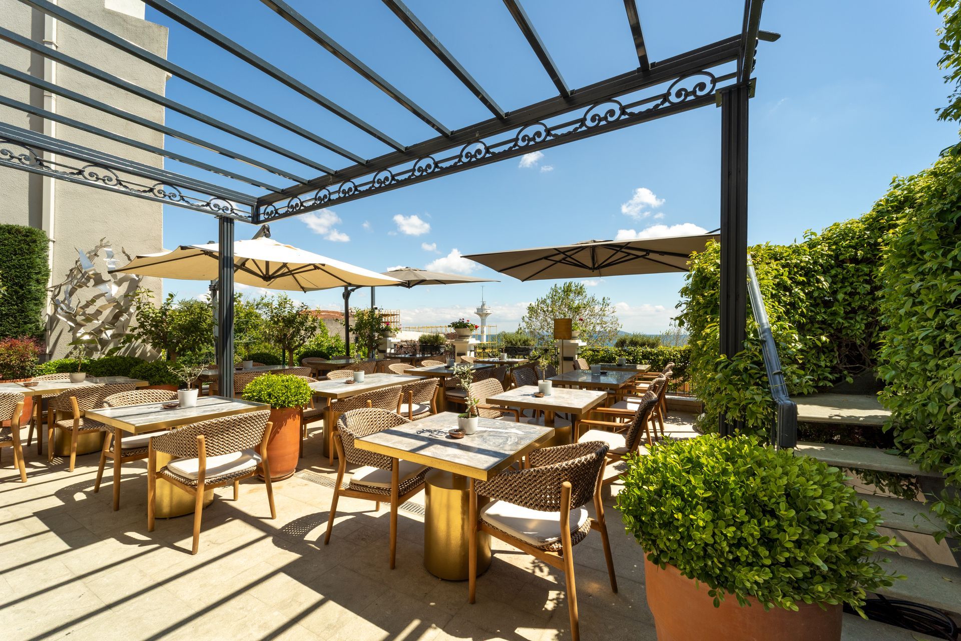 Outdoor restaurant terrace with wooden tables, potted plants, and shade sails overlooking the sea