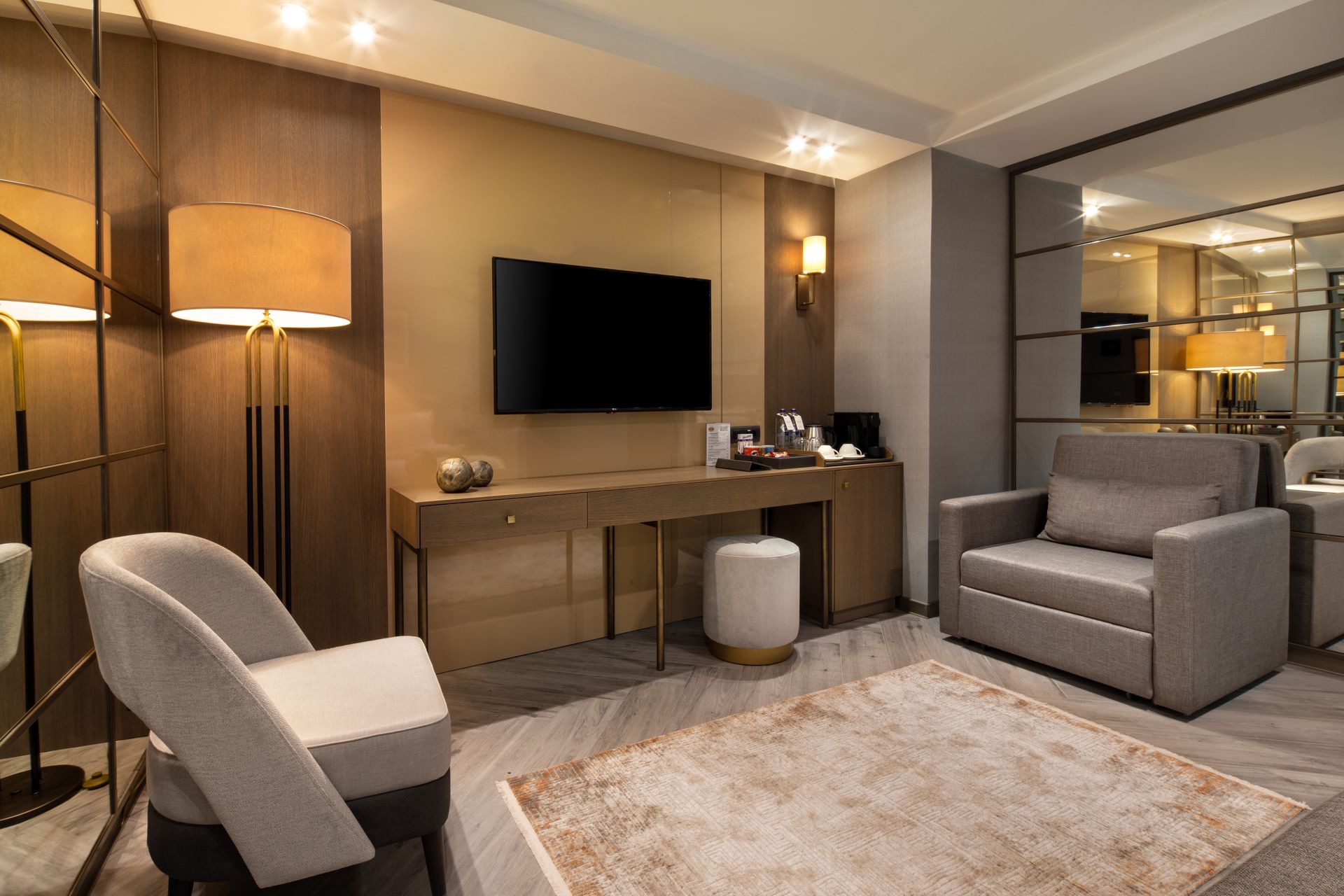 Modern hotel room with a wall-mounted TV, desk, lamp, and two gray chairs on a carpeted floor.