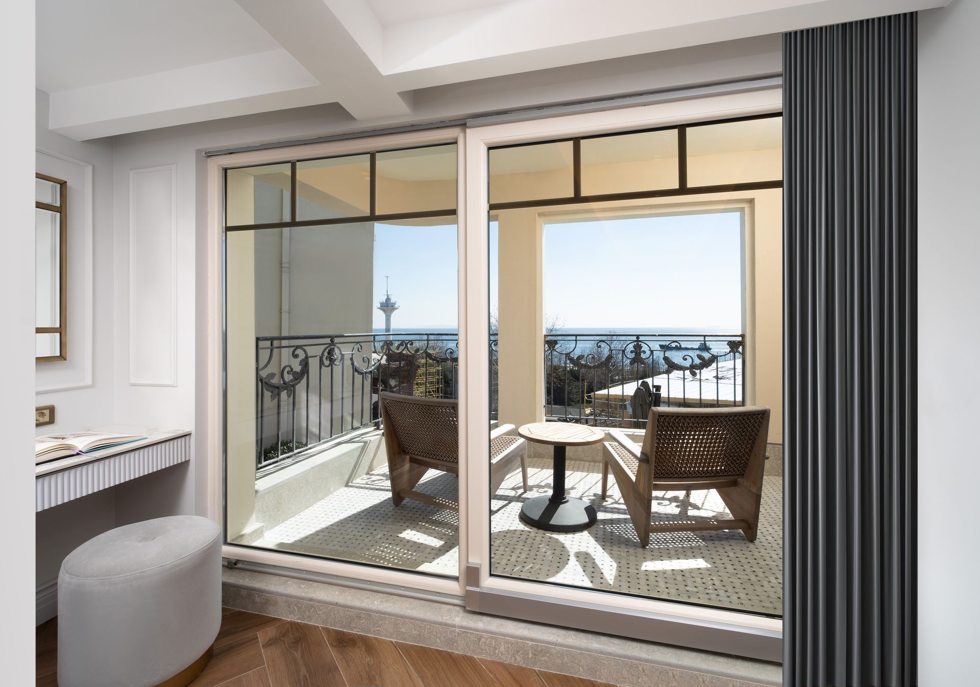 Bright room opening to a balcony with chairs, a table, and ocean view beyond the railing.