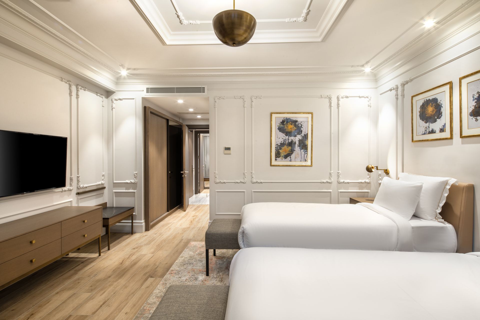 Elegant white hotel room with twin beds, TV, wood floor, and framed wall art.