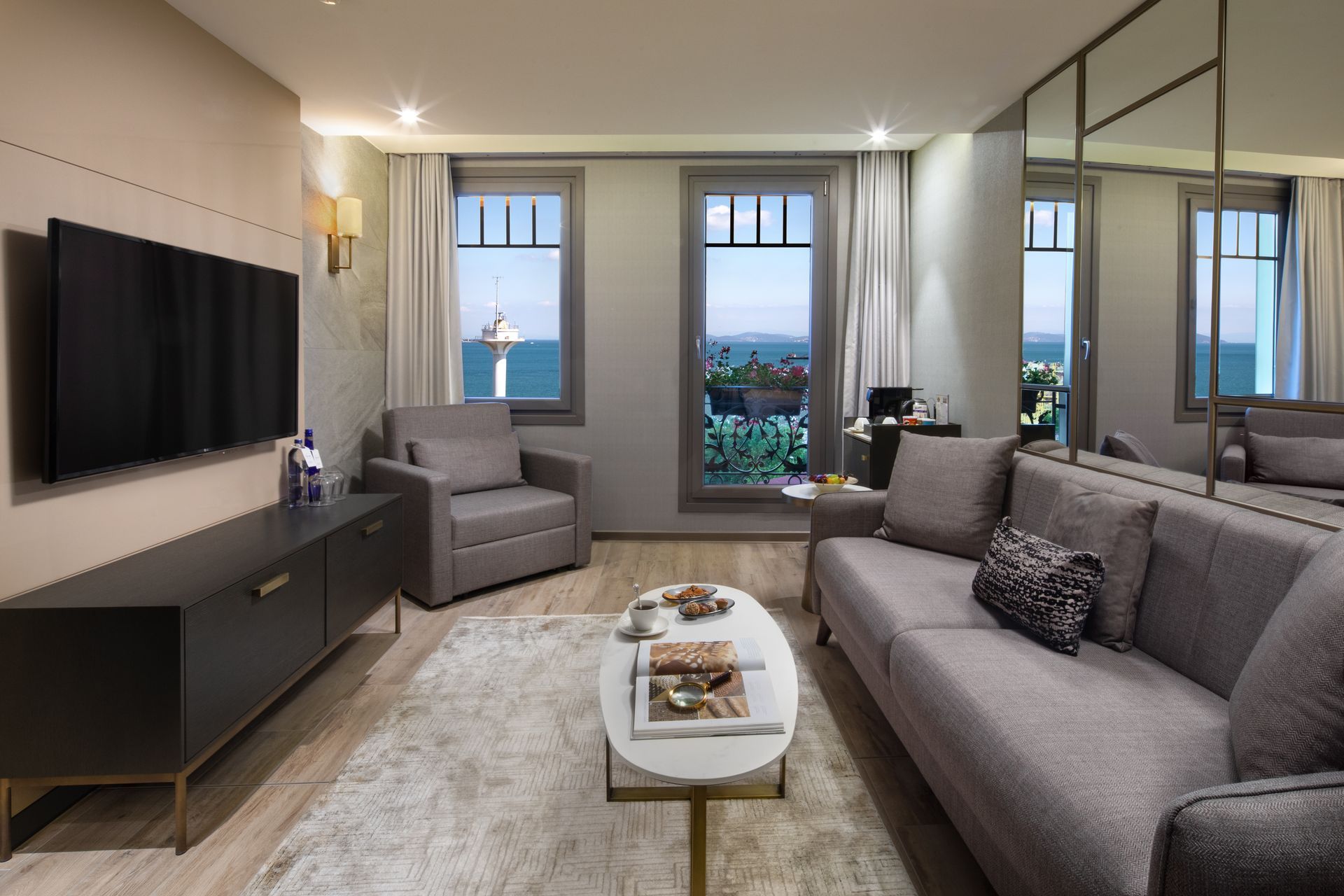 Modern living room with gray sofa, armchair, wall-mounted TV, coffee table, and large windows overlooking greenery