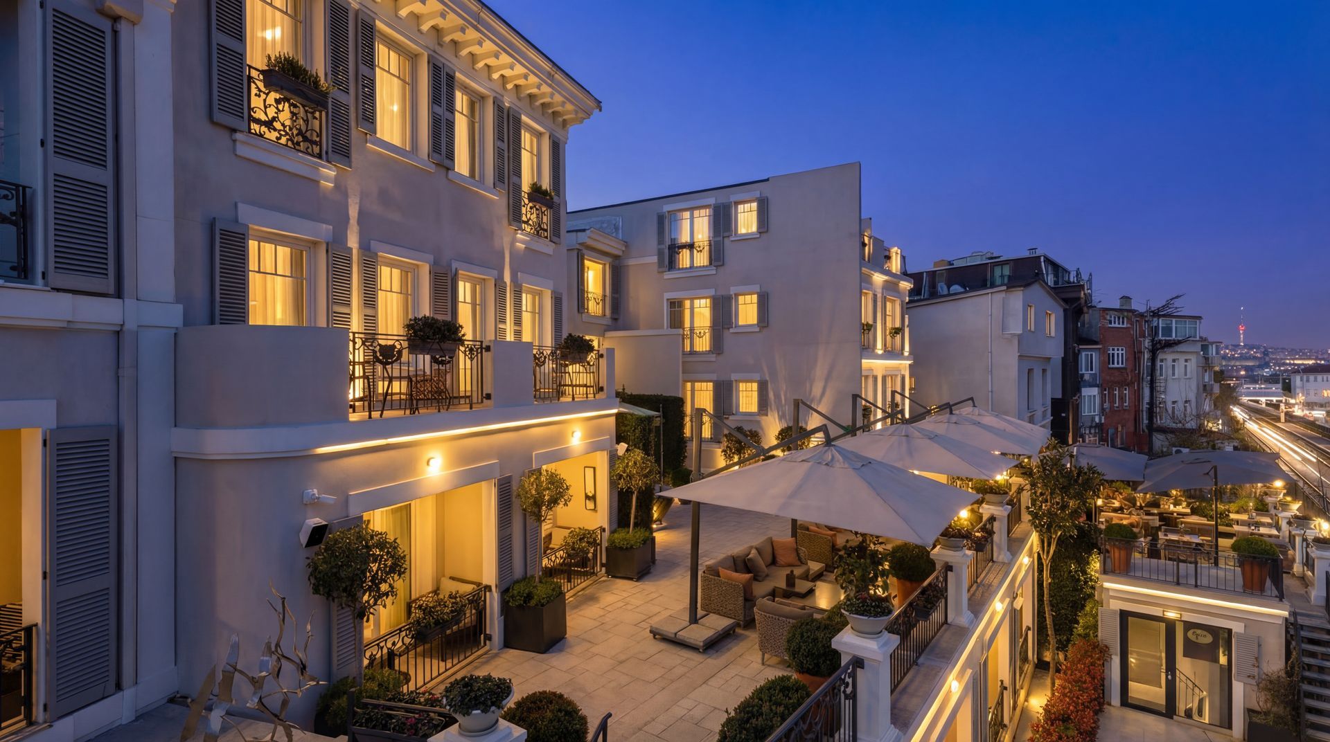 Dusk view of a modern apartment complex with lit balconies, courtyard trees, and a street lined with buildings