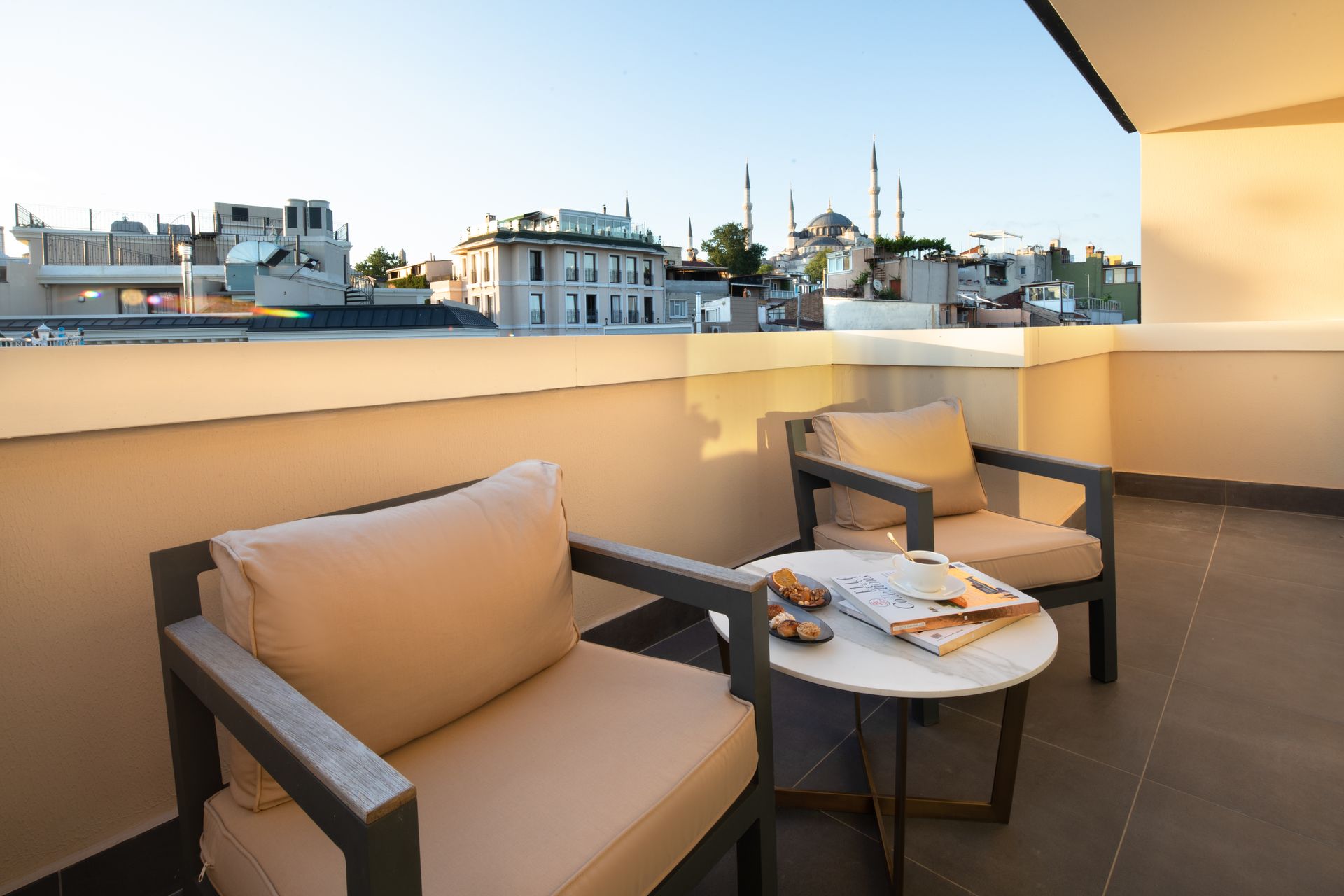 Balcony with two beige chairs and a small table overlooking a city at sunset