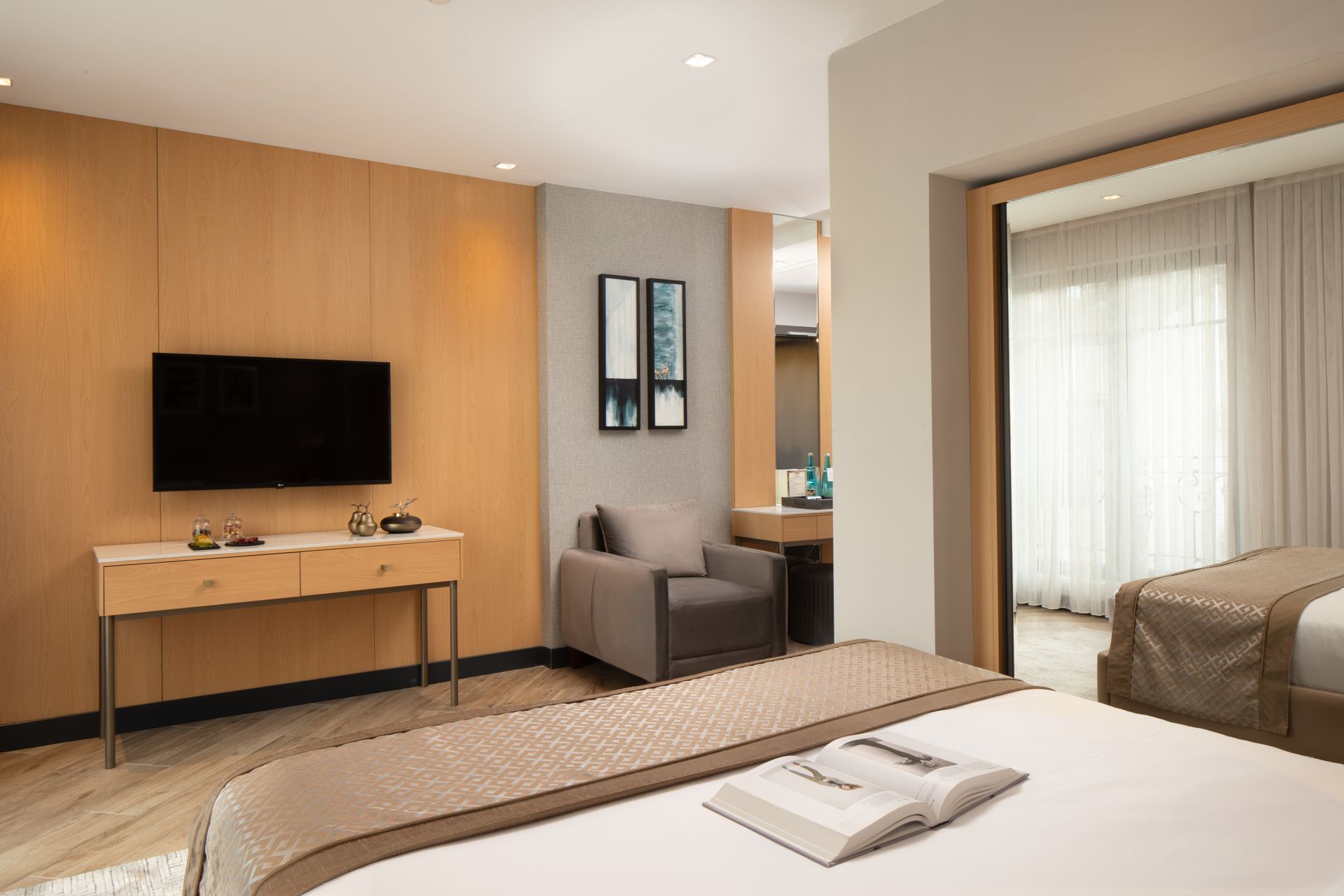 Modern hotel room with bed, wall-mounted TV, desk, armchair, and large window with sheer curtains