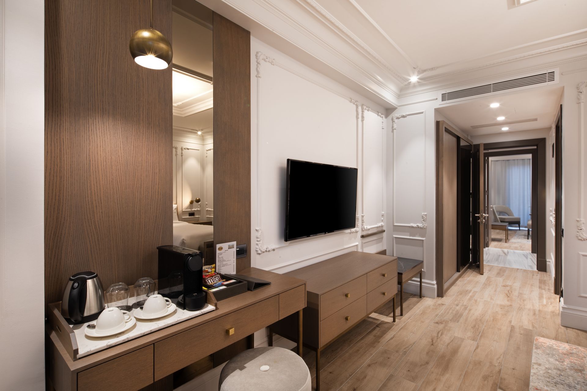 Modern hotel room with a wall-mounted TV, desk, and hallway view in neutral tones