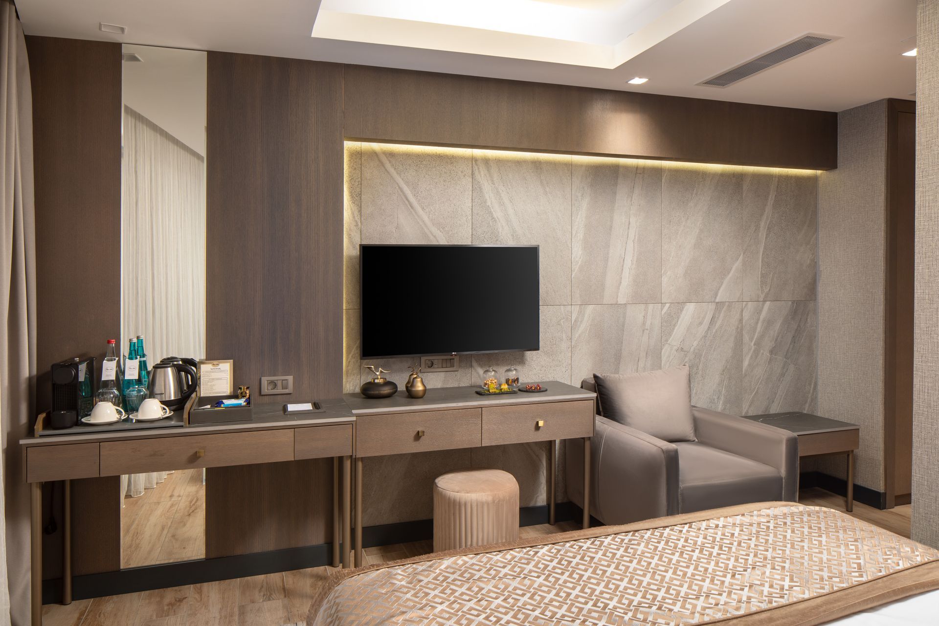 Modern hotel room with desk, TV, armchair, and bed in a beige and wood-toned interior | Mula Hotel