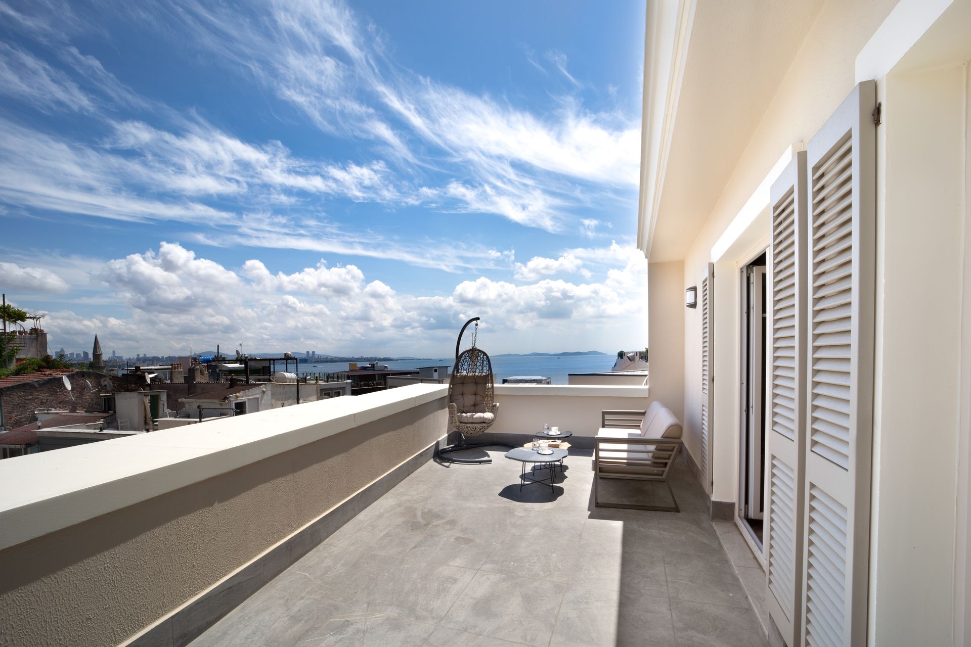 Sunlit rooftop terrace with lounge chairs, ocean view, and white shuttered doors on the right