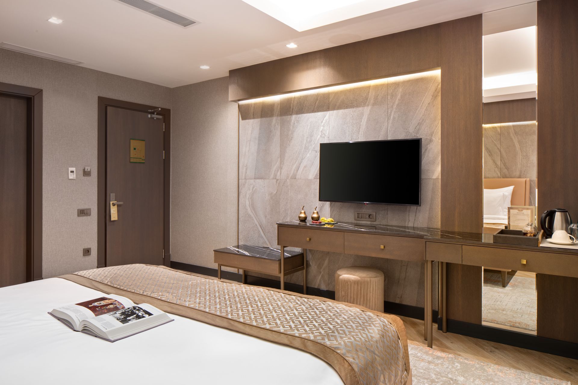 Modern hotel room with a bed, TV, desk, mirror, and warm lighting.