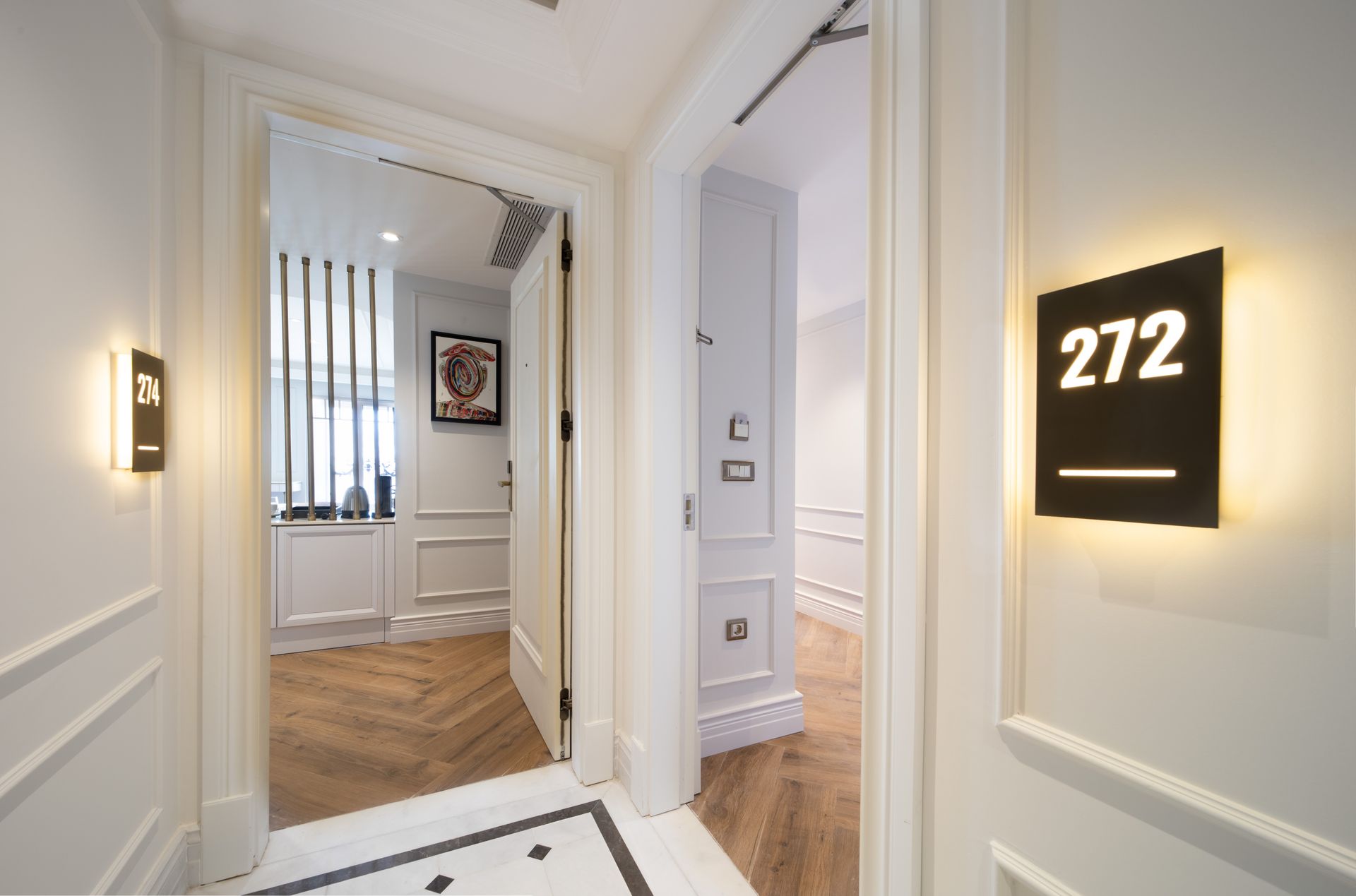 Bright hallway with white walls, wooden floor, and black room number sign 272 beside an open doorway.