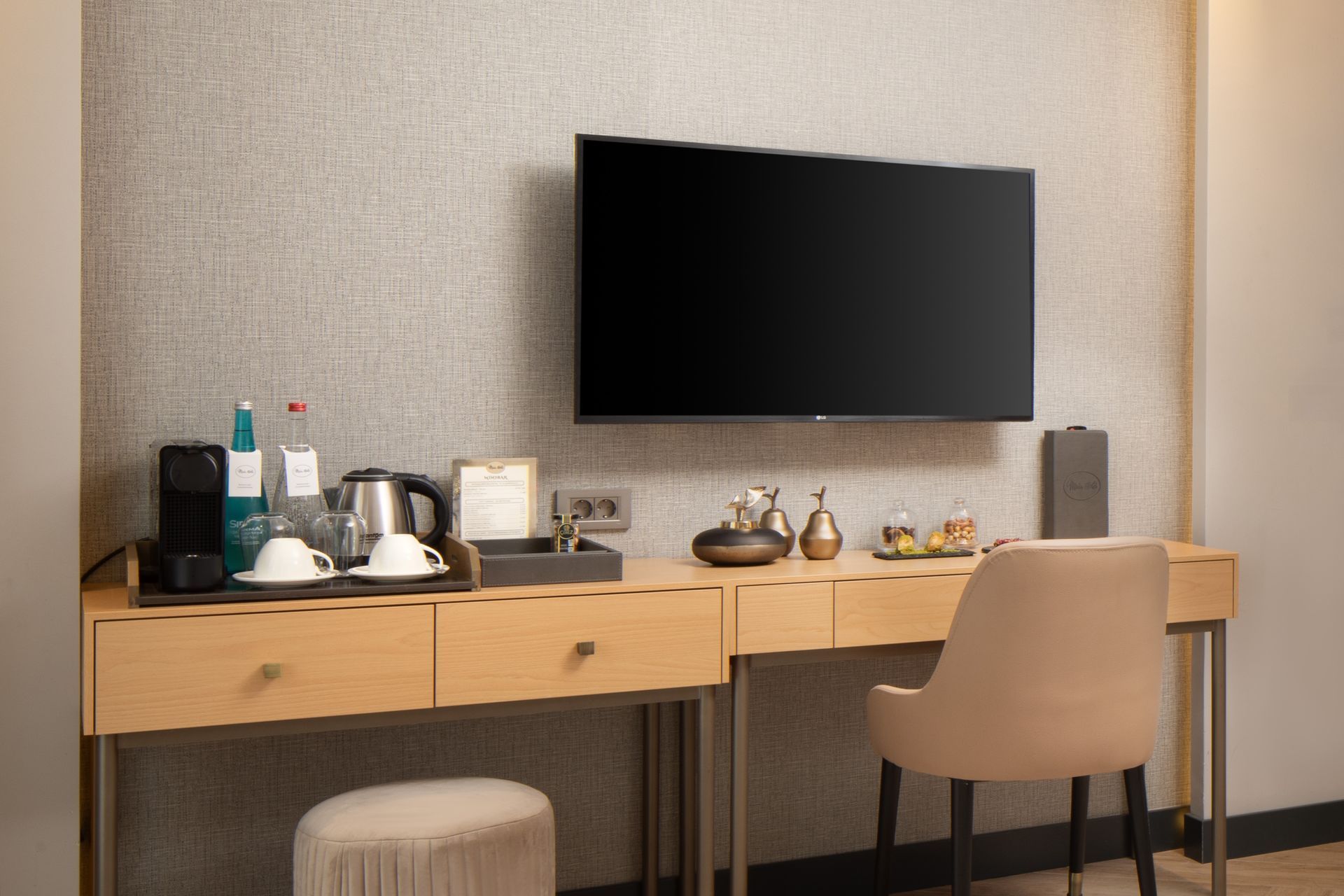 Modern hotel desk with TV, lamp, kettle, and chair against a textured wall