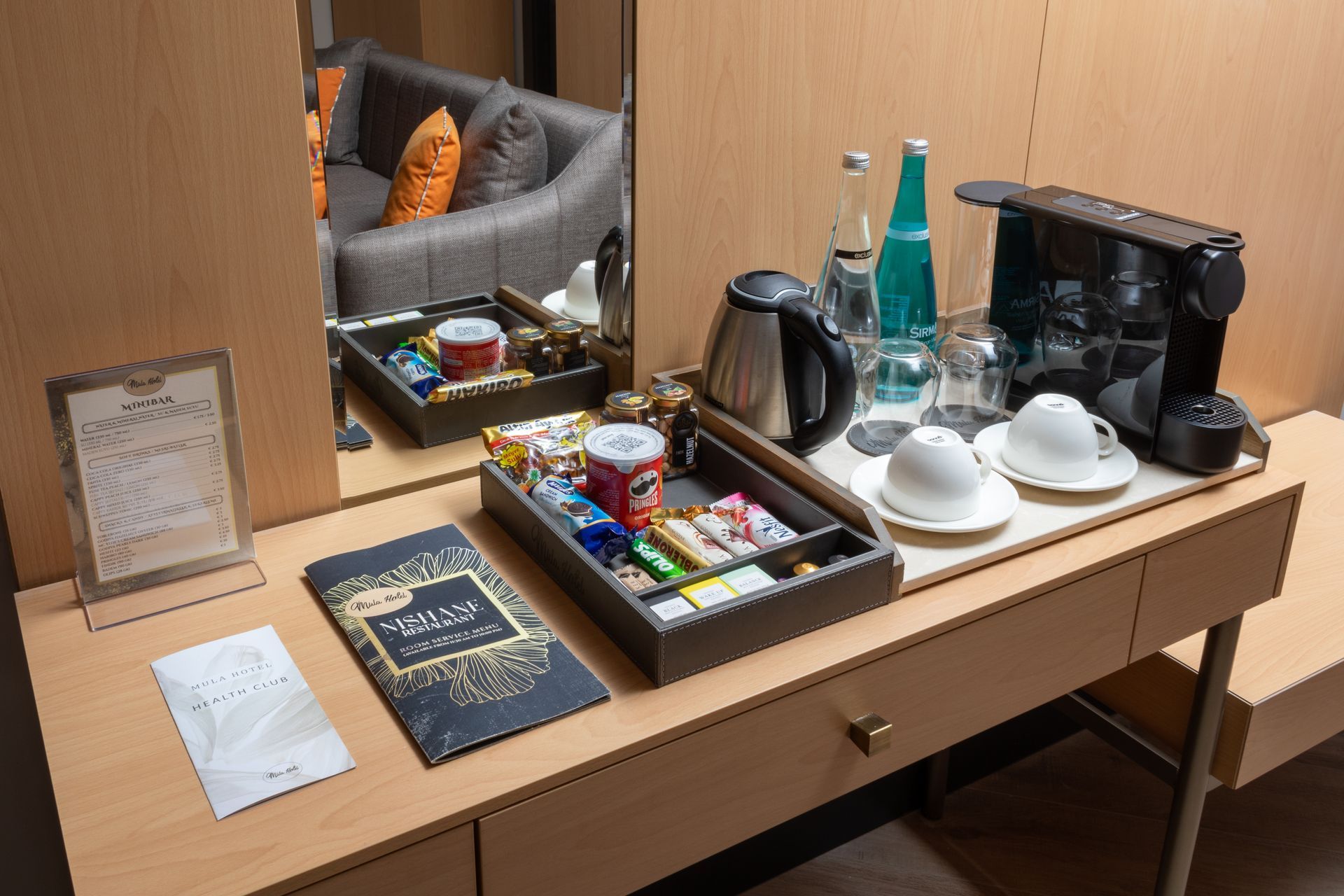 Hotel room coffee and tea tray on a desk with cups, kettle, bottles, snacks, and a mirror. | Mula Hotel