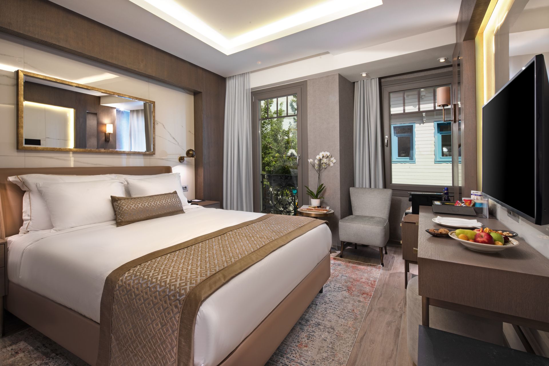 Modern hotel room with a large bed, armchair, window view, and desk under warm lighting