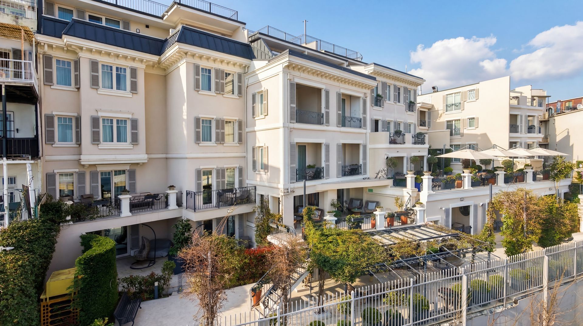 Modern beige apartment buildings with balconies and greenery along a fenced walkway in daylight